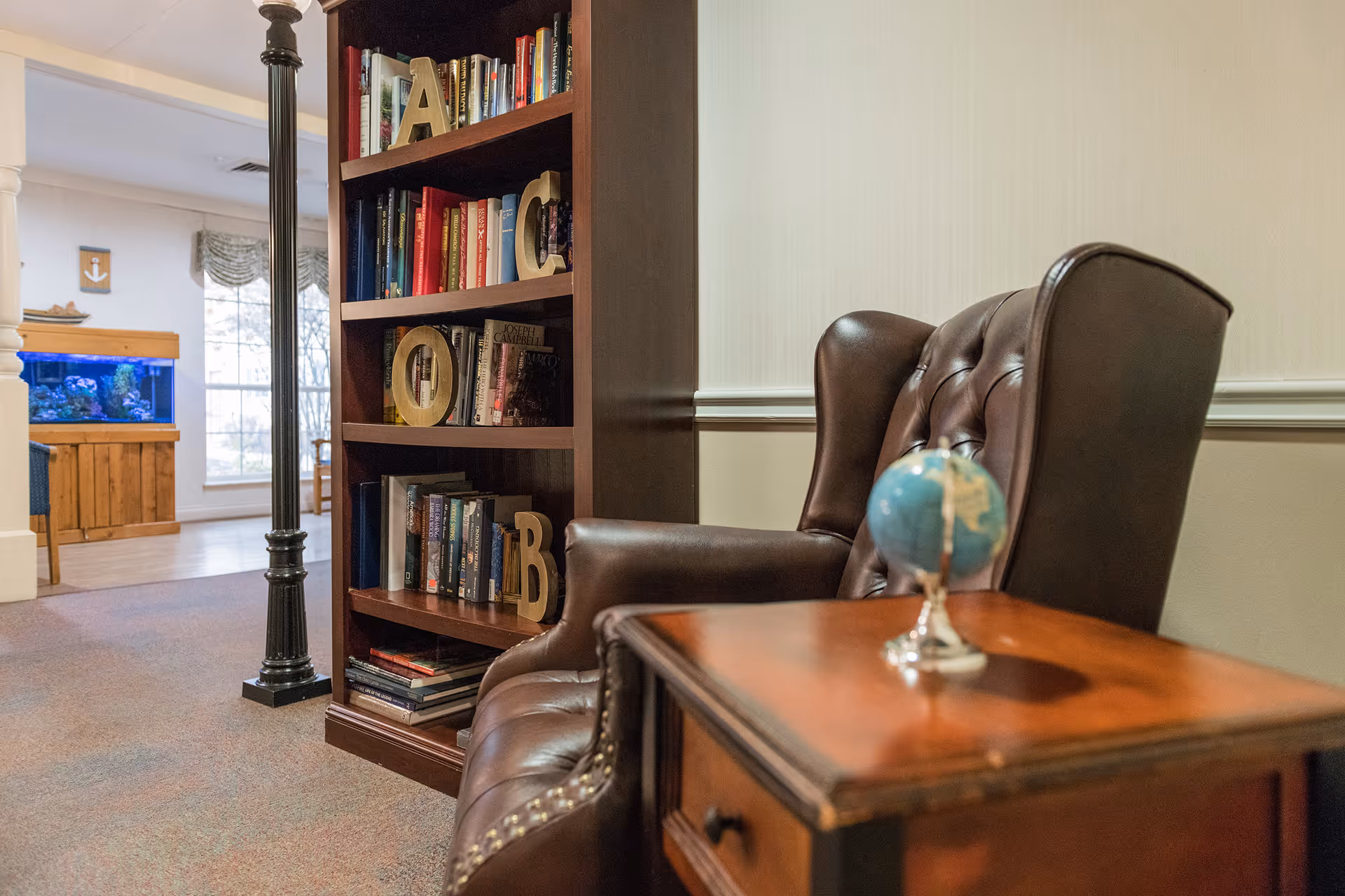 Cozy common-room scene with a leather armchair, wooden side table holding a small globe, and a bookshelf with books and decorative letters.