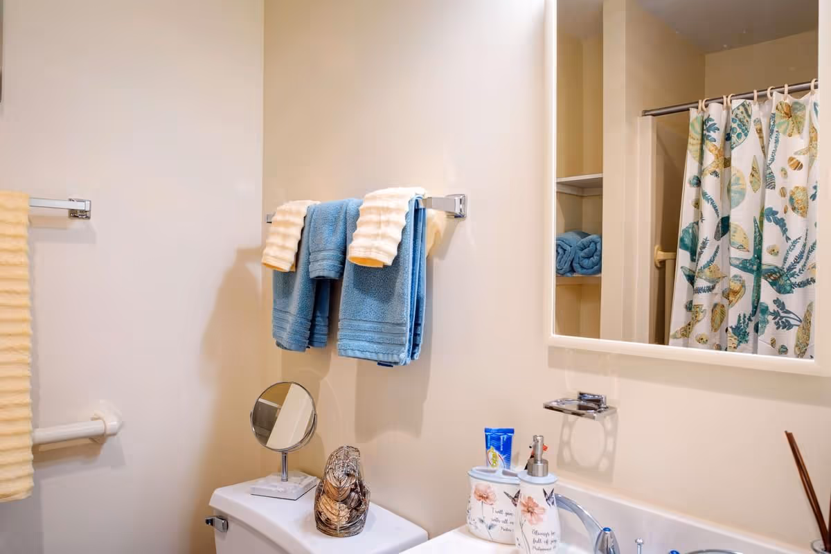 A bathroom with beige walls featuring a white toilet topped with a small round mirror and decorative item. Above the toilet, there are two blue towels with two cream-colored towels hanging on a towel rack. A sink with floral soap dispensers and a toothbrush holder is visible, along with a mirror reflecting a shower curtain with a leaf and shell pattern. Shelves with rolled blue towels are also seen in the reflection.