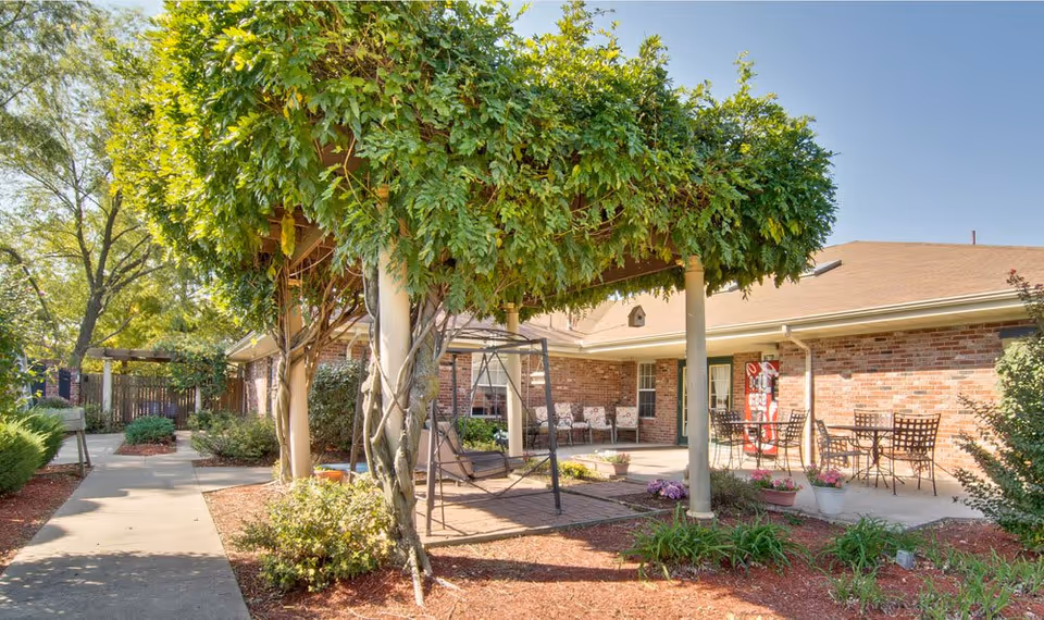 Outdoor courtyard with a vine-covered pergola, patio swing and tables in front of a brick senior-living building.