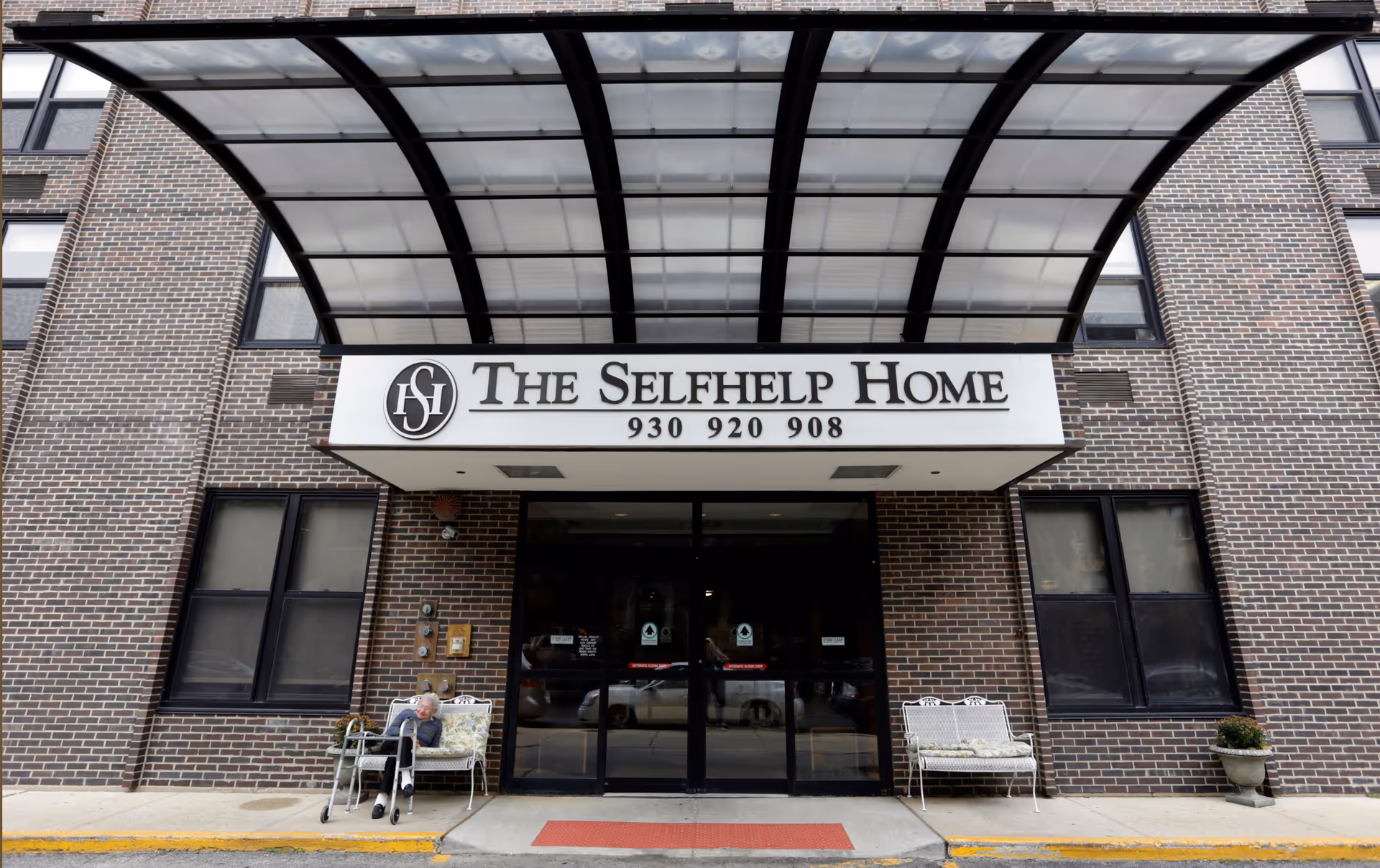 Entrance of The Selfhelp Home Senior Living Community with a large awning above the doorway. Two benches are placed on either side of the entrance, with an elderly person sitting on the left bench using a walker. The building exterior is made of brown brick.