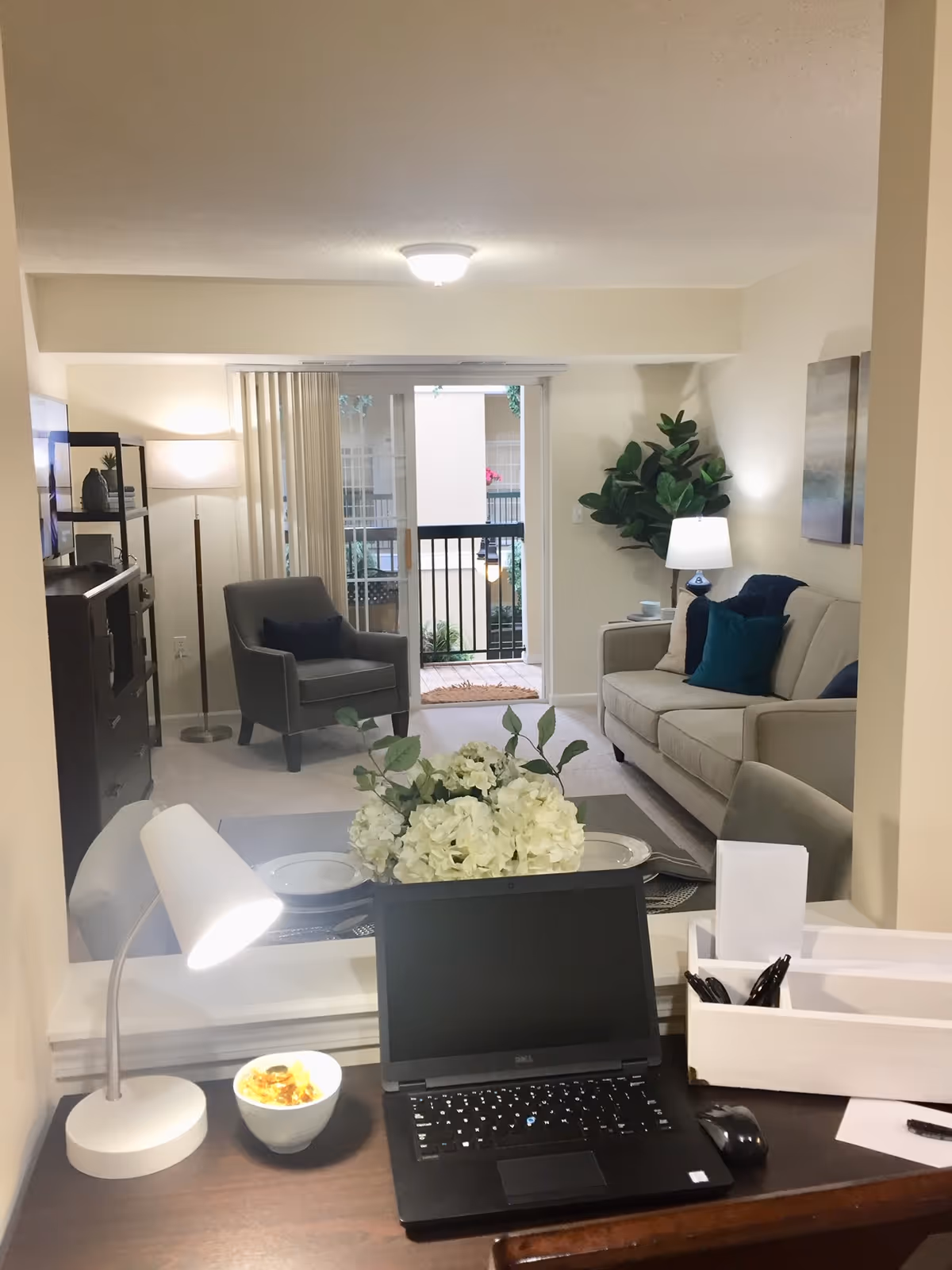 A cozy living room with a beige sofa adorned with blue and green pillows, a gray armchair, a floor lamp, and a TV on a stand. There is a sliding glass door leading to a balcony with a railing and some plants outside. In the foreground, there is a desk with a laptop, a white desk lamp, a bowl of snacks, and a white organizer with pens and papers. A bouquet of white flowers is placed on the coffee table in the living room.