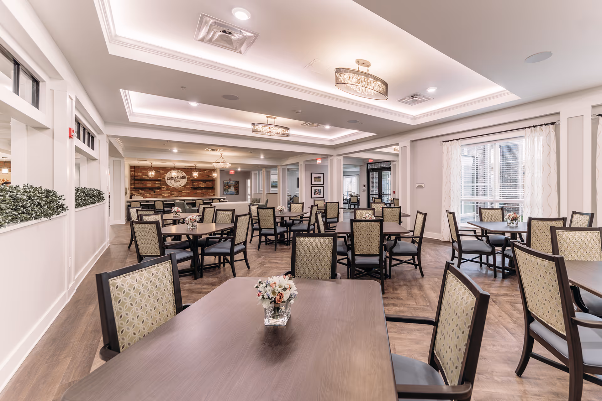 A spacious and well-lit dining room with multiple tables and chairs arranged neatly. Each table has a small floral centerpiece. The room features large windows with sheer curtains allowing natural light to fill the space. The ceiling has recessed lighting and decorative light fixtures. The walls are painted white with some decorative plants along a half wall on the left side.