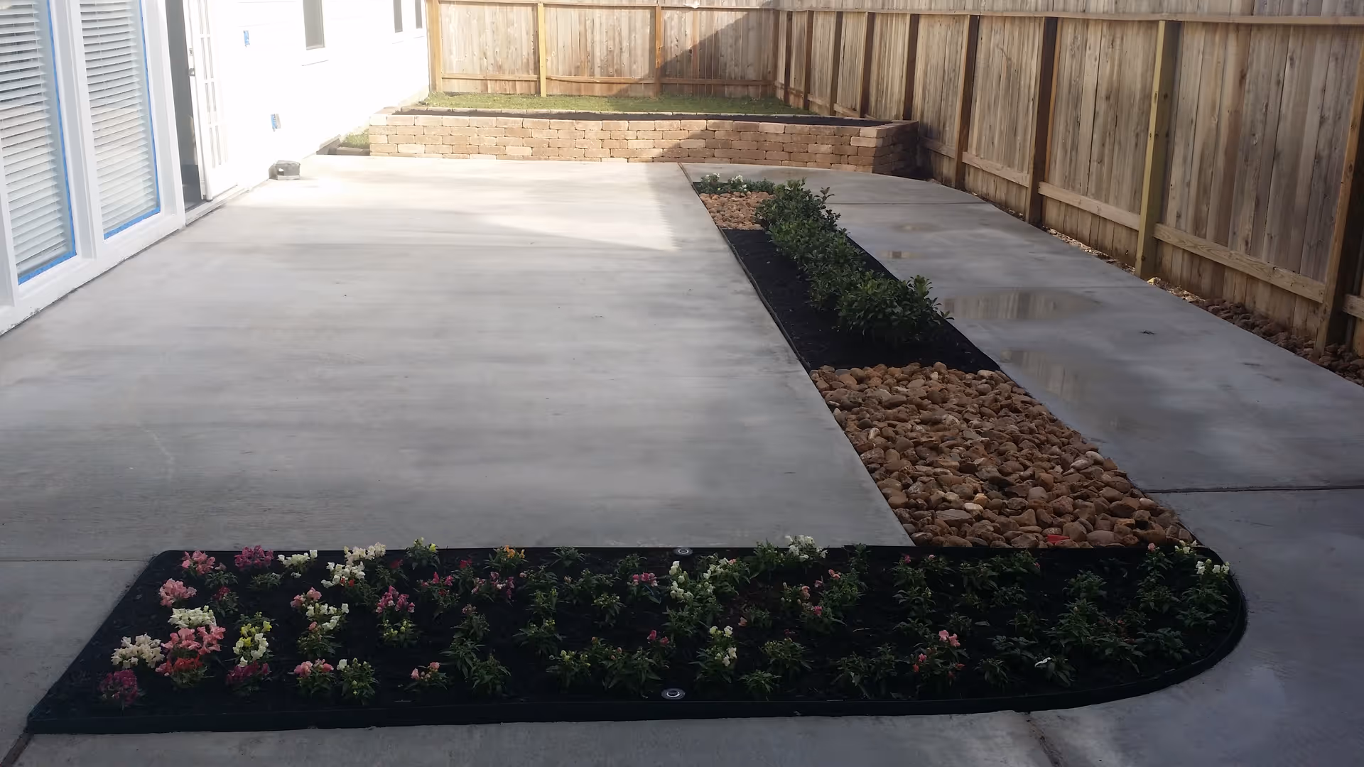 A fenced outdoor patio area with a concrete floor, a narrow garden bed with small green plants and flowers, and a section filled with rocks. A raised grassy area with a retaining wall is visible in the background.