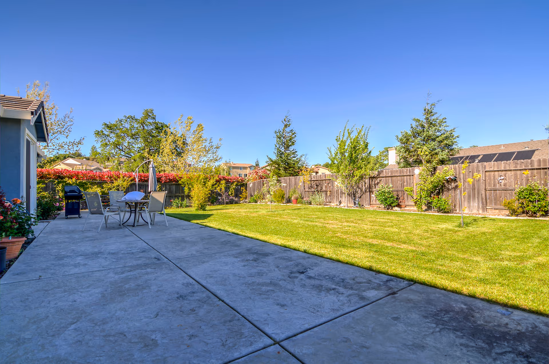 Concrete patio with an outdoor dining table, umbrella and grill next to a large fenced grassy backyard under a clear blue sky.