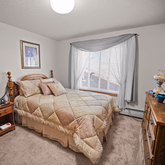 A cozy bedroom with a wooden bed frame and a beige and light brown patchwork quilt. The bed has several pillows and is positioned next to a window with white blinds and sheer white and gray curtains. There is a wooden nightstand with a phone and a remote control, and a wooden dresser with a vintage-style lamp and some small items on top. The room has beige carpet and light-colored walls with a framed picture hanging above the bed.