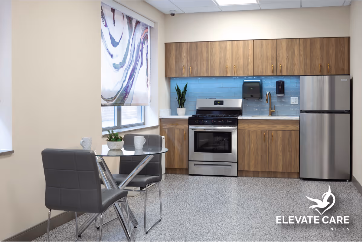 A modern kitchen area with wooden cabinets, a stainless steel stove and refrigerator, a blue tile backsplash, and a small round glass table with two gray chairs. There is a window with a decorative blind and a small plant on the countertop.