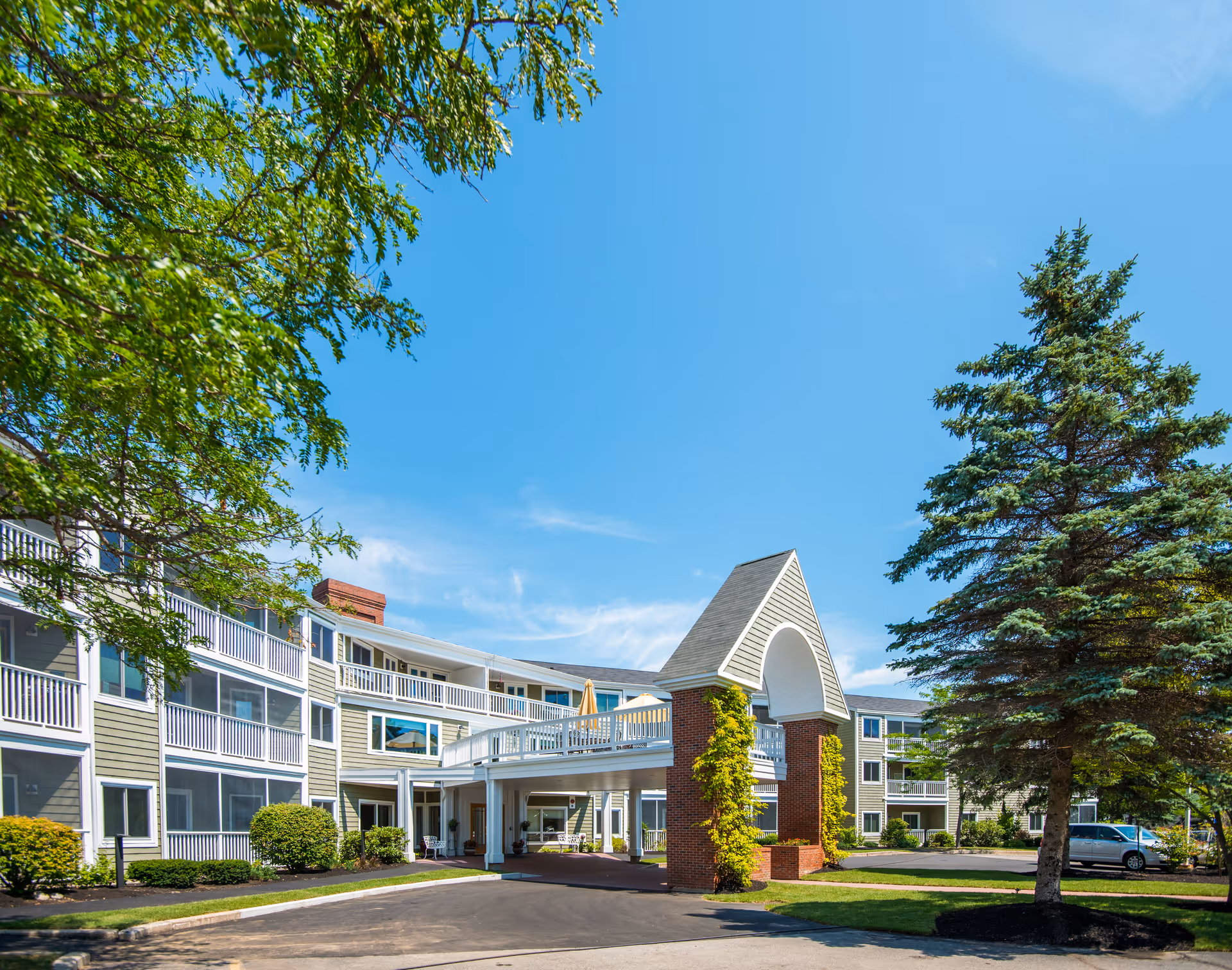 Front exterior of a multi-story senior living building with a covered entrance, balconies, trees, and landscaping under a blue sky.