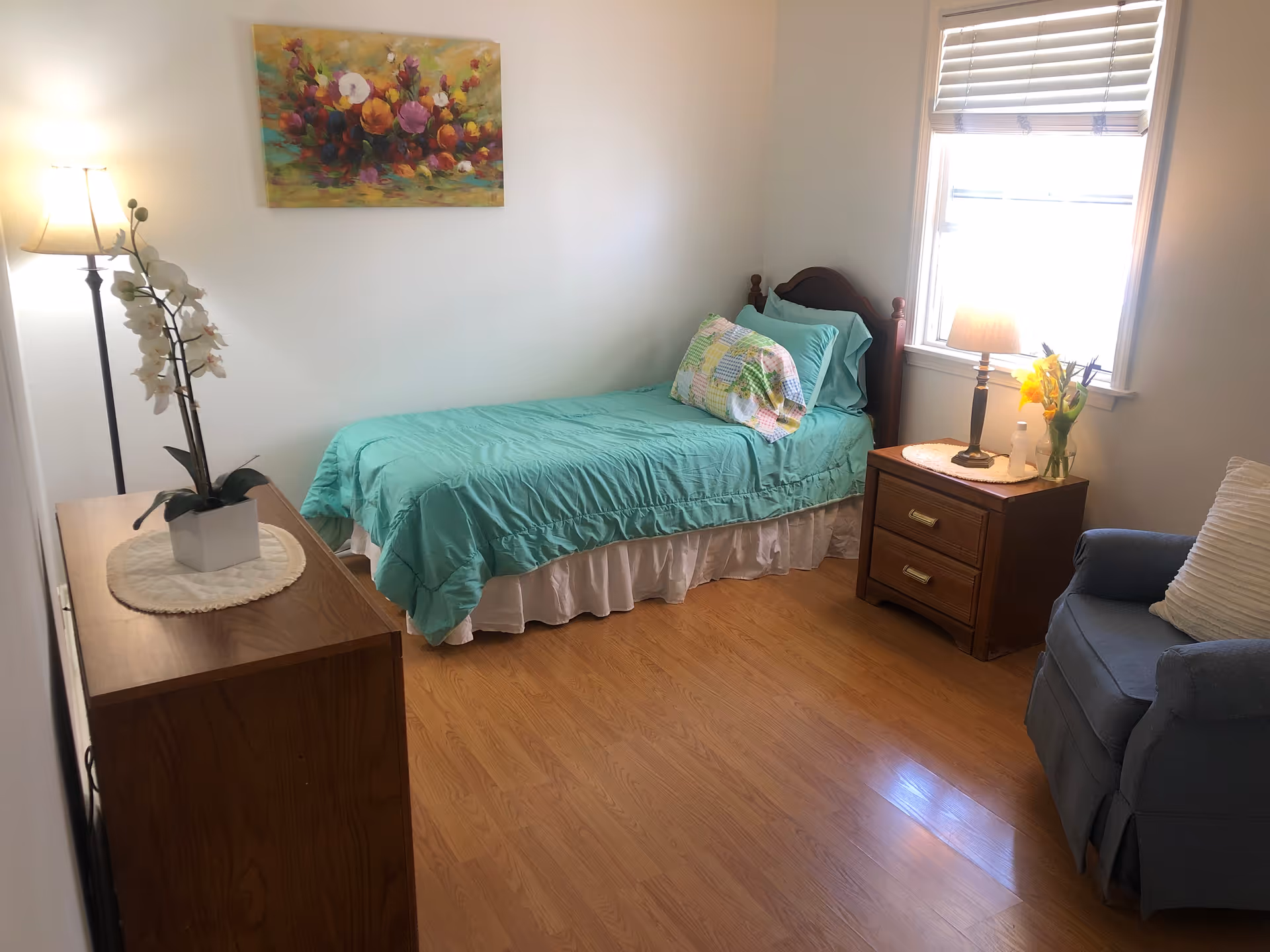 A small, cozy bedroom with a single bed covered in a turquoise bedspread and a colorful pillow. There is a wooden nightstand with a lamp and a vase of flowers next to the bed, a blue armchair with a white pillow, a wooden dresser with a white orchid plant, and a floor lamp. A colorful floral painting hangs on the wall above the bed, and a window with blinds lets in natural light.