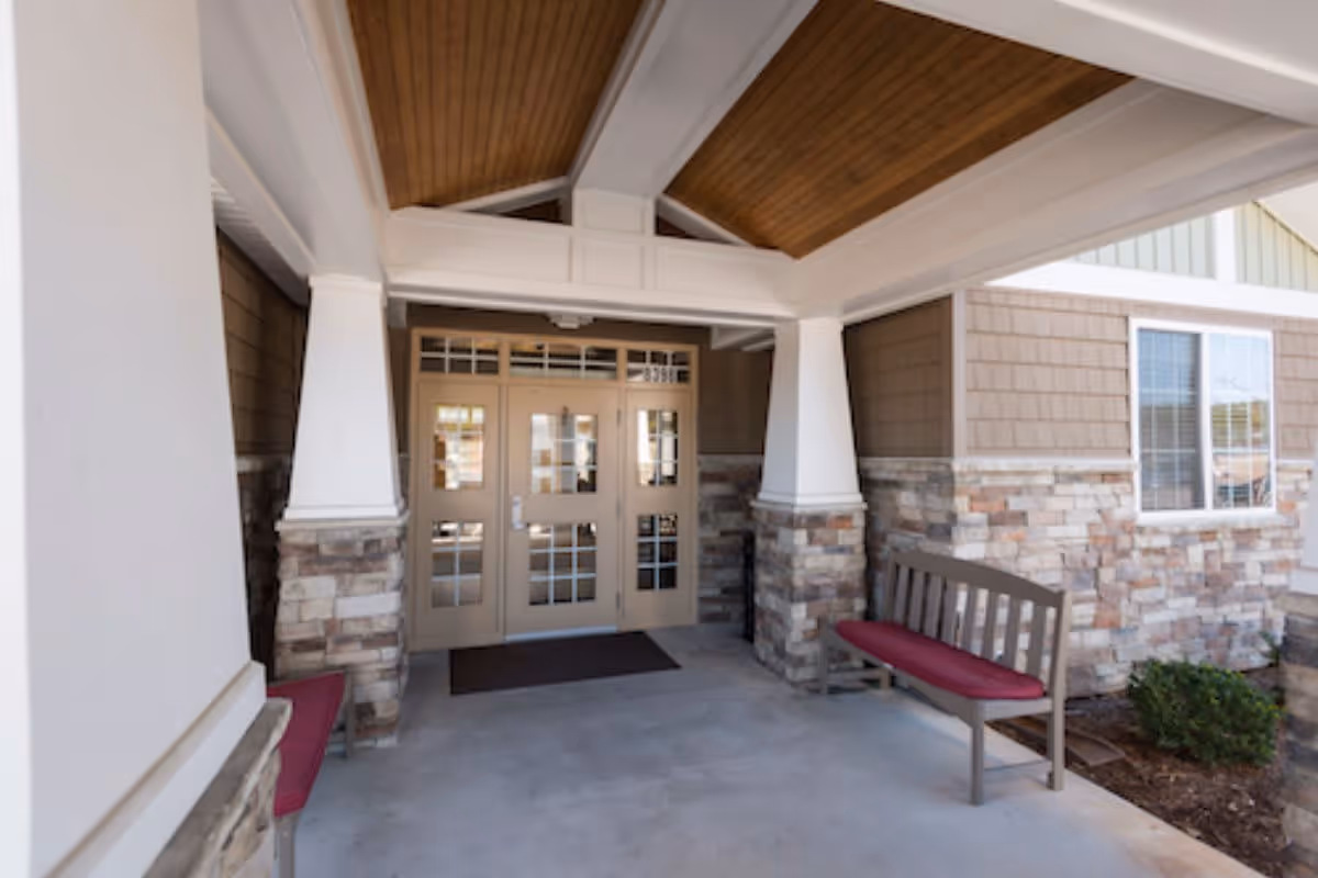 Covered entrance area of a building with double glass doors, stone and wood siding, two benches with red cushions on either side, and a wooden ceiling.
