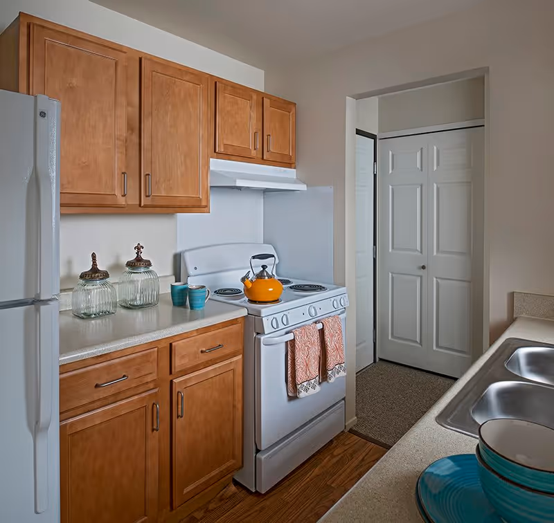 A kitchen area with wooden cabinets, a white refrigerator, a white stove with an orange kettle on top, two orange patterned towels hanging on the oven door, two glass jars and two blue mugs on the countertop, and a double sink with stacked bowls and plates nearby. There is a doorway with white double doors in the background.