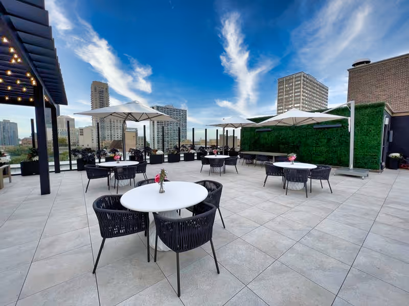 Outdoor rooftop patio area with round white tables and black chairs, each table decorated with a small vase of flowers. Large white umbrellas provide shade, and a green wall is visible on one side. The city skyline with tall buildings is seen in the background under a blue sky with some clouds.