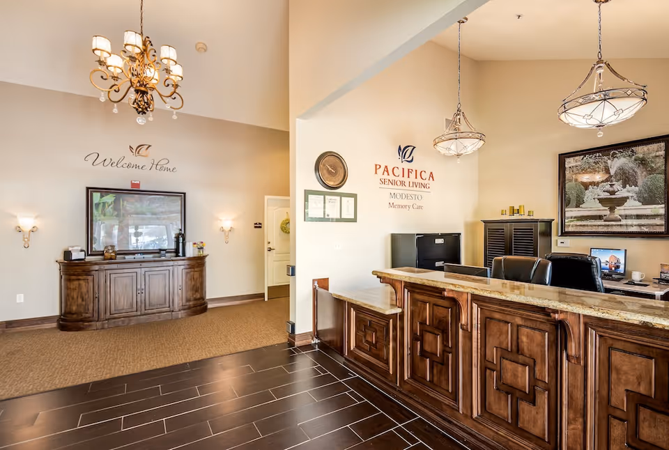Reception area of a senior living facility with a wooden front desk, two hanging light fixtures, and a wall sign that reads 'PACIFICA SENIOR LIVING MODESTO Memory Care'. To the left, there is a wooden cabinet with a framed picture above it and a wall decal that says 'Welcome Home'. The space has beige walls, dark wood flooring near the desk, and carpet in the adjacent area.