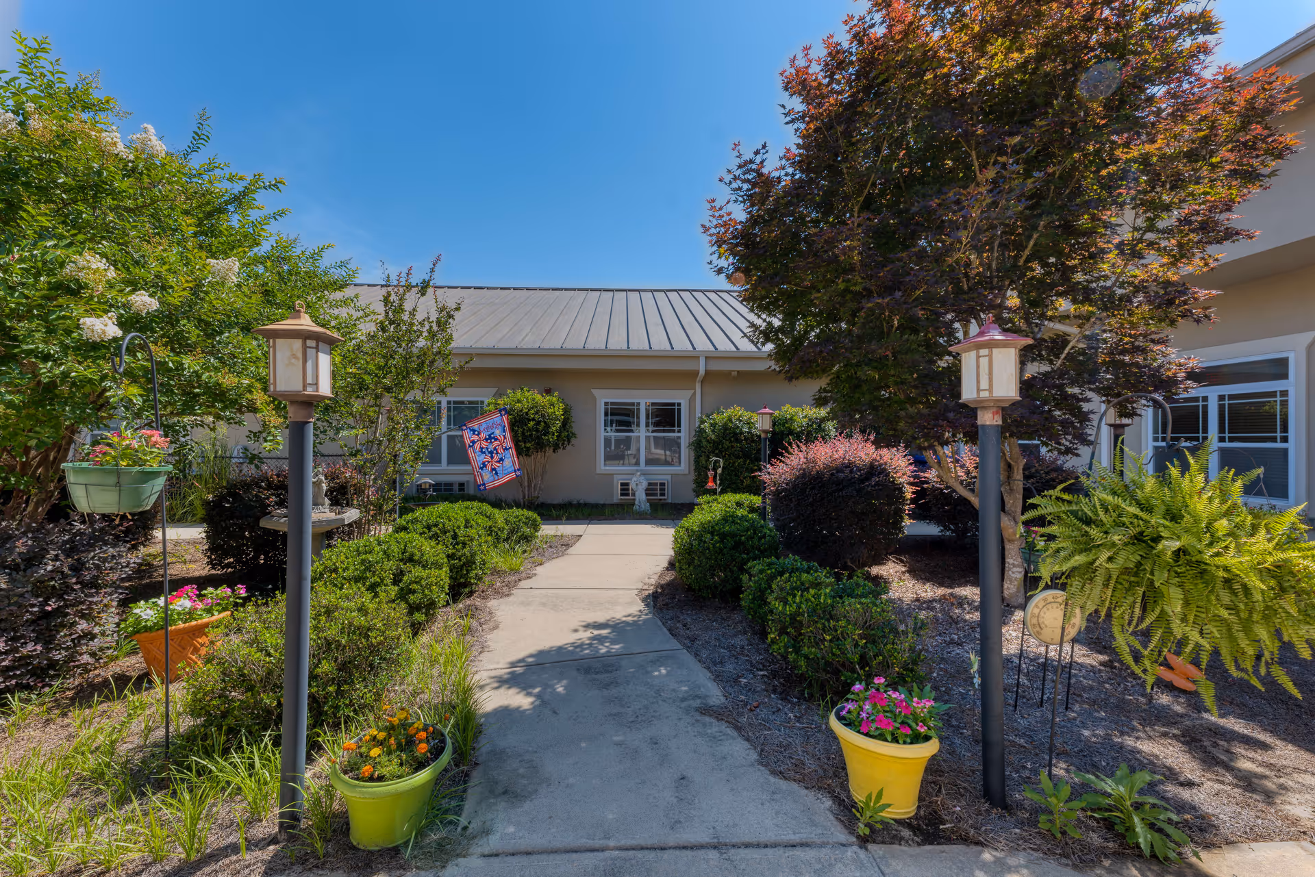 A sunny outdoor garden area with a concrete walkway leading to a building entrance. The garden is landscaped with various green shrubs, flowering plants in pots, and small trees. There are two lamp posts on either side of the walkway and a decorative flag hanging near the building. The sky is clear and blue.