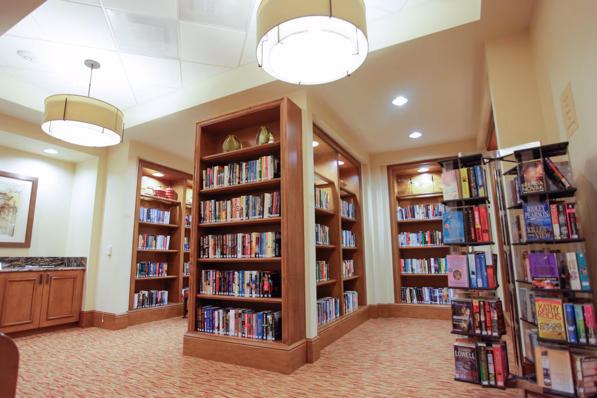 Interior view of a library or reading room with wooden bookshelves filled with books. The room has warm lighting with two large ceiling lamps, carpeted floor, and a small cabinet with a granite countertop on the left side. There is also a rotating book display rack on the right side.