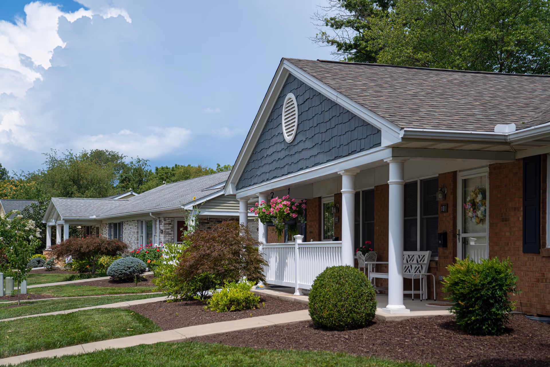 Exterior view of a single-story residential building with a covered porch, white columns, and well-maintained landscaping including bushes and hanging flower baskets under a partly cloudy sky.