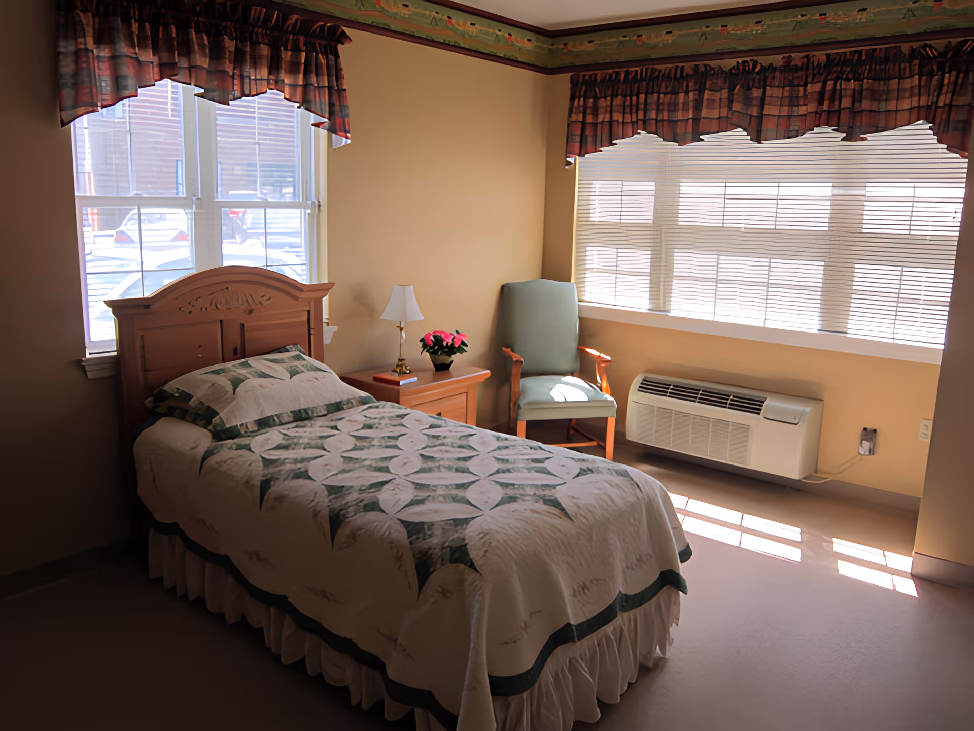 A sunlit single bedroom with a bed dressed in a patterned quilt, a nightstand and lamp, and a chair by large windows.