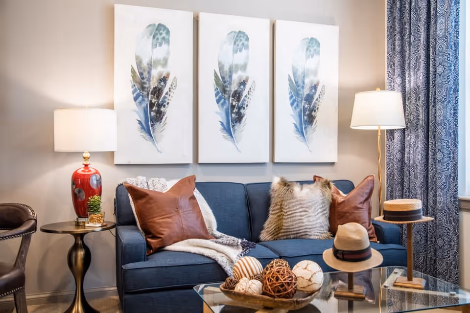 A cozy living room with a blue sofa adorned with brown and furry pillows, a throw blanket, and a glass coffee table holding decorative balls and a hat. On the wall above the sofa are three vertical paintings of blue and green feathers. There are two lamps, one with a red base on a small round side table to the left, and a tall floor lamp to the right near a window with blue patterned curtains.