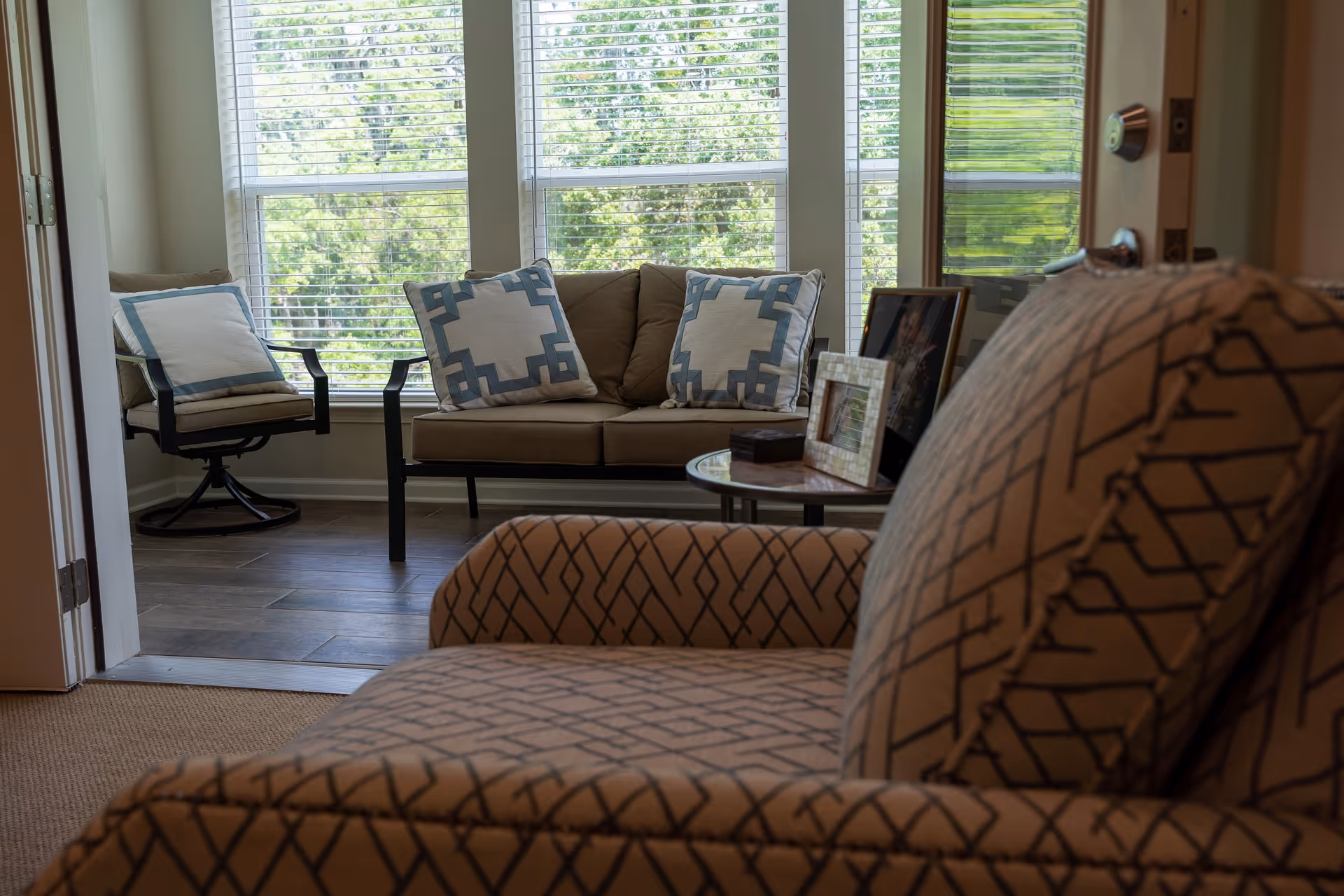 A cozy living room area with a patterned armchair in the foreground, a small round table with framed photos, and a beige sofa with two decorative pillows featuring a geometric design. Large windows with white blinds allow natural light to fill the room, and a cushioned chair with a pillow is positioned near the windows.