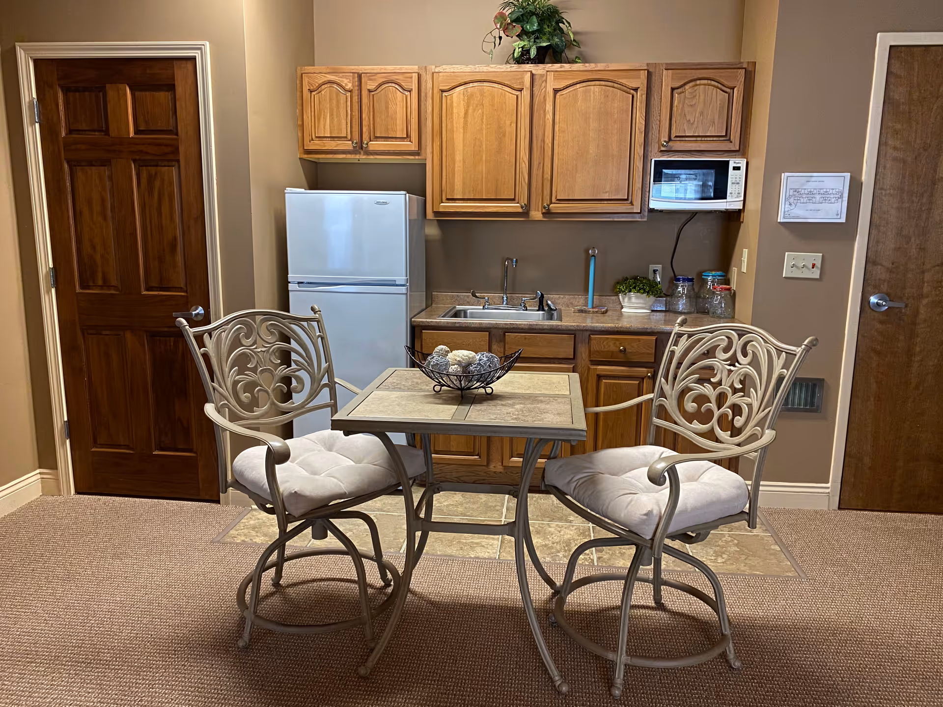 Small kitchen area with wooden cabinets, a white refrigerator, a microwave mounted above the counter, and a sink. In front of the kitchen is a small square table with a decorative bowl on top and two cushioned metal chairs with ornate backs. The room has beige walls and carpeted flooring with a tiled section in front of the kitchen. There are two wooden doors visible on either side of the kitchen.