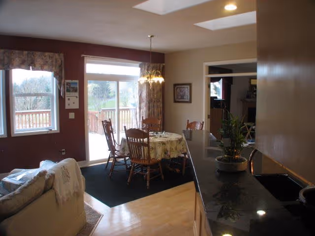 Bright open dining area with a round table and wooden chairs next to sliding glass doors, adjacent to a granite-topped kitchen counter and a sofa.