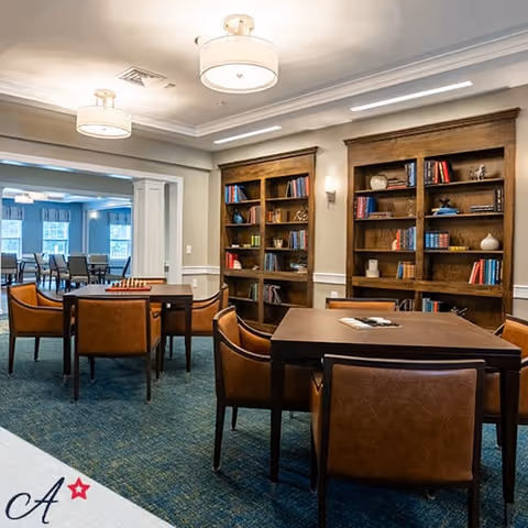A cozy interior room with wooden bookshelves filled with books and decorative items, several square tables surrounded by brown leather chairs, and ceiling lights. In the background, another room with more tables and chairs is visible.