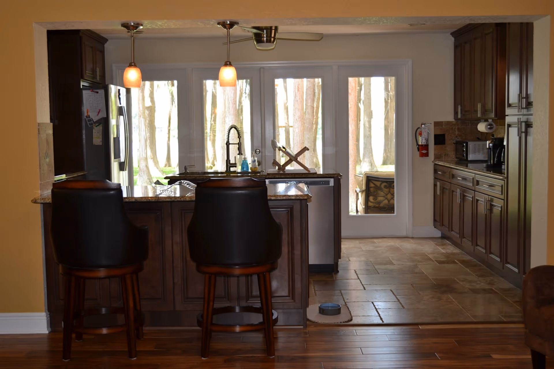 Open kitchen with a central island, two bar stools, pendant lights, and large glass doors showing trees outside.