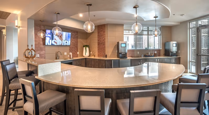 Modern communal kitchen and curved breakfast bar with pendant lights, stools, a wall-mounted TV, and beverage stations.