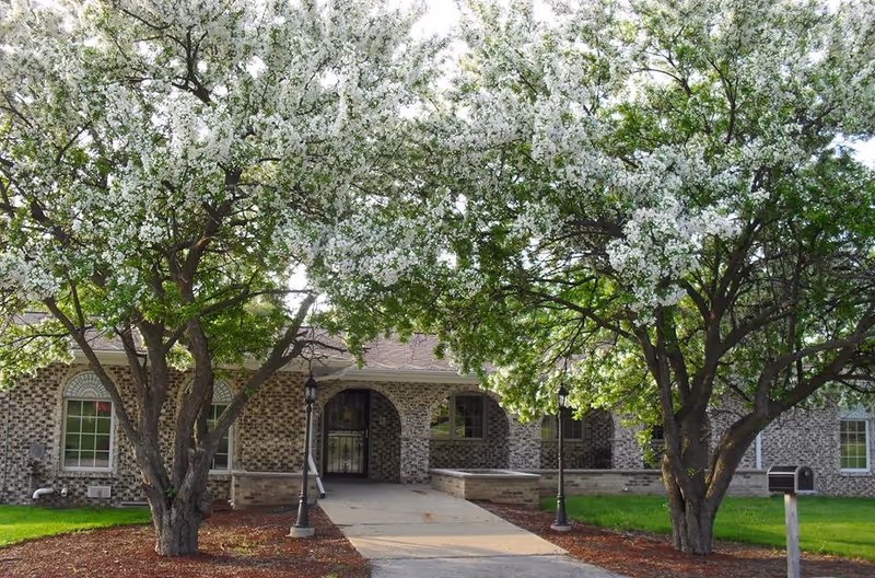 Front exterior view of a brick building with arched doorways and windows, flanked by two large trees with white blossoms, a concrete walkway leading to the entrance, and green grass on either side.