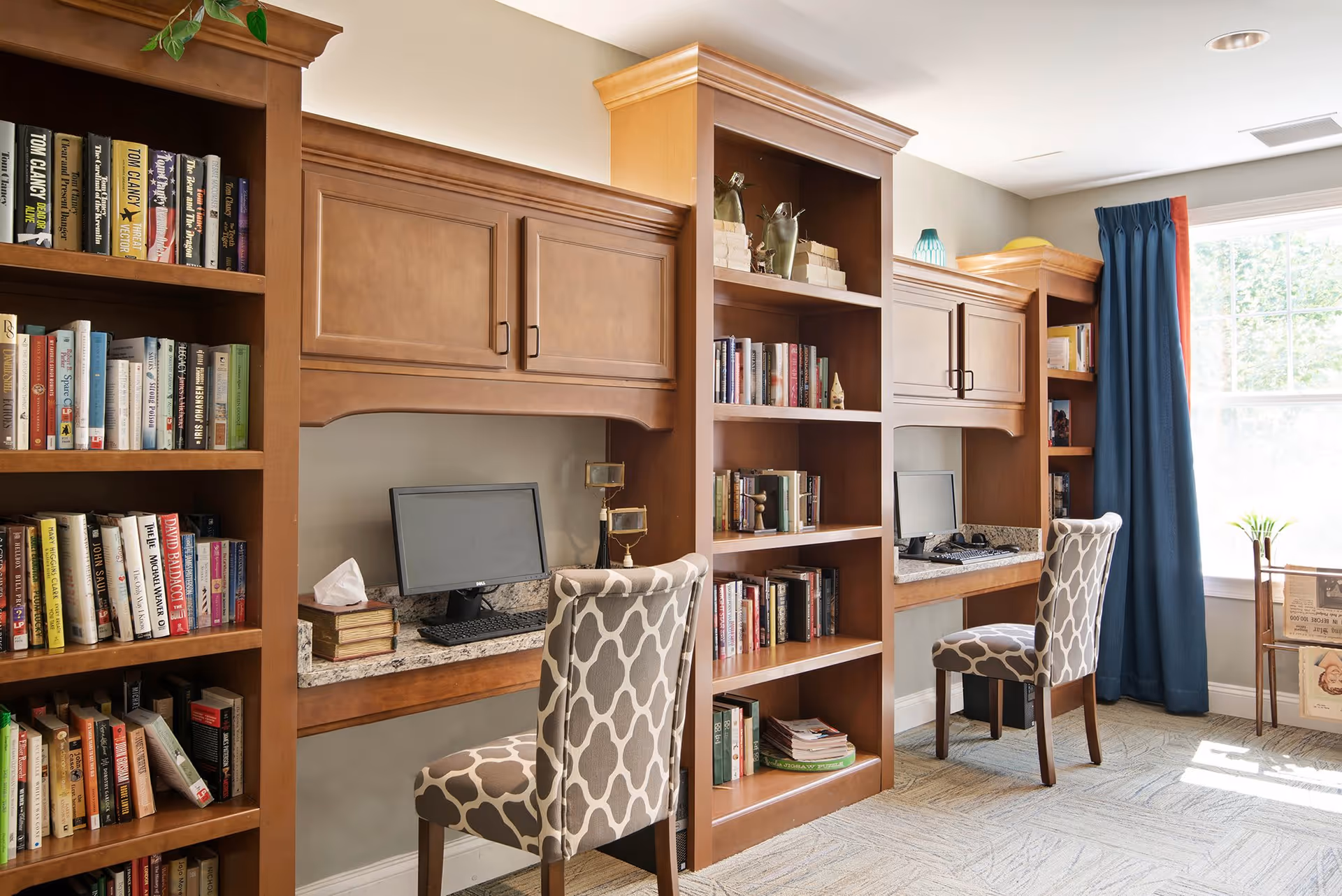 A cozy study area in a senior living facility with built-in wooden bookshelves filled with books, two computer workstations with monitors and patterned chairs, and a large window with blue curtains letting in natural light.