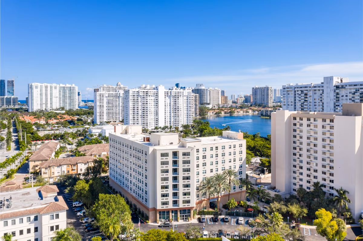 Aerial exterior view of The Sterling Aventura building among nearby high-rise residences with a waterfront and clear blue sky.