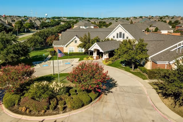 Aerial view of The Auberge at Valley Ranch senior living facility showing a circular driveway with landscaped bushes and trees, an American flag on a flagpole, and a large building with a pitched roof surrounded by greenery and neighboring houses in the background.