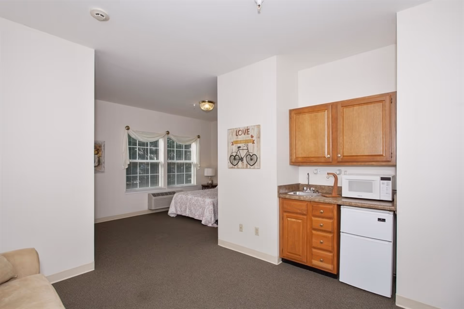 Studio-style room with a small kitchenette on the right and a bed near windows in the background.