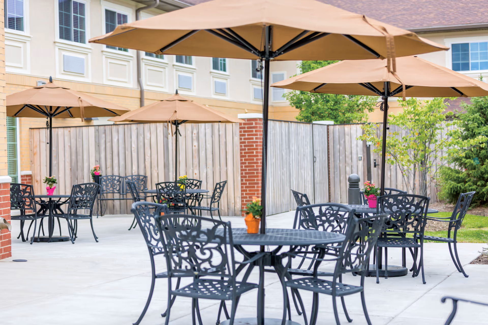 Outdoor patio area with several black metal tables and chairs, each table shaded by large beige umbrellas. Small potted plants with colorful flowers are placed on the tables. The patio is surrounded by a wooden fence and part of a building with multiple windows is visible in the background.