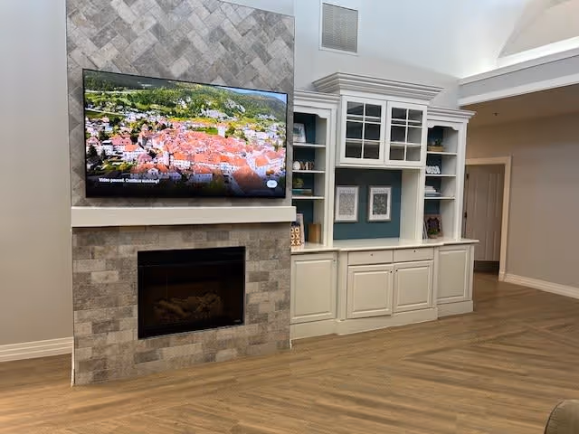 A cozy interior living space featuring a wall-mounted flat-screen TV above a stone fireplace. To the right, there is a built-in white cabinet with glass doors and open shelves displaying decorative items. The room has wood flooring and neutral-colored walls with a doorway leading to another area.