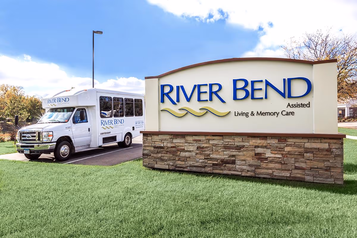 A large stone-based sign reading 'RIVER BEND Assisted Living & Memory Care' beside a parked River Bend shuttle bus on a grassy lawn.