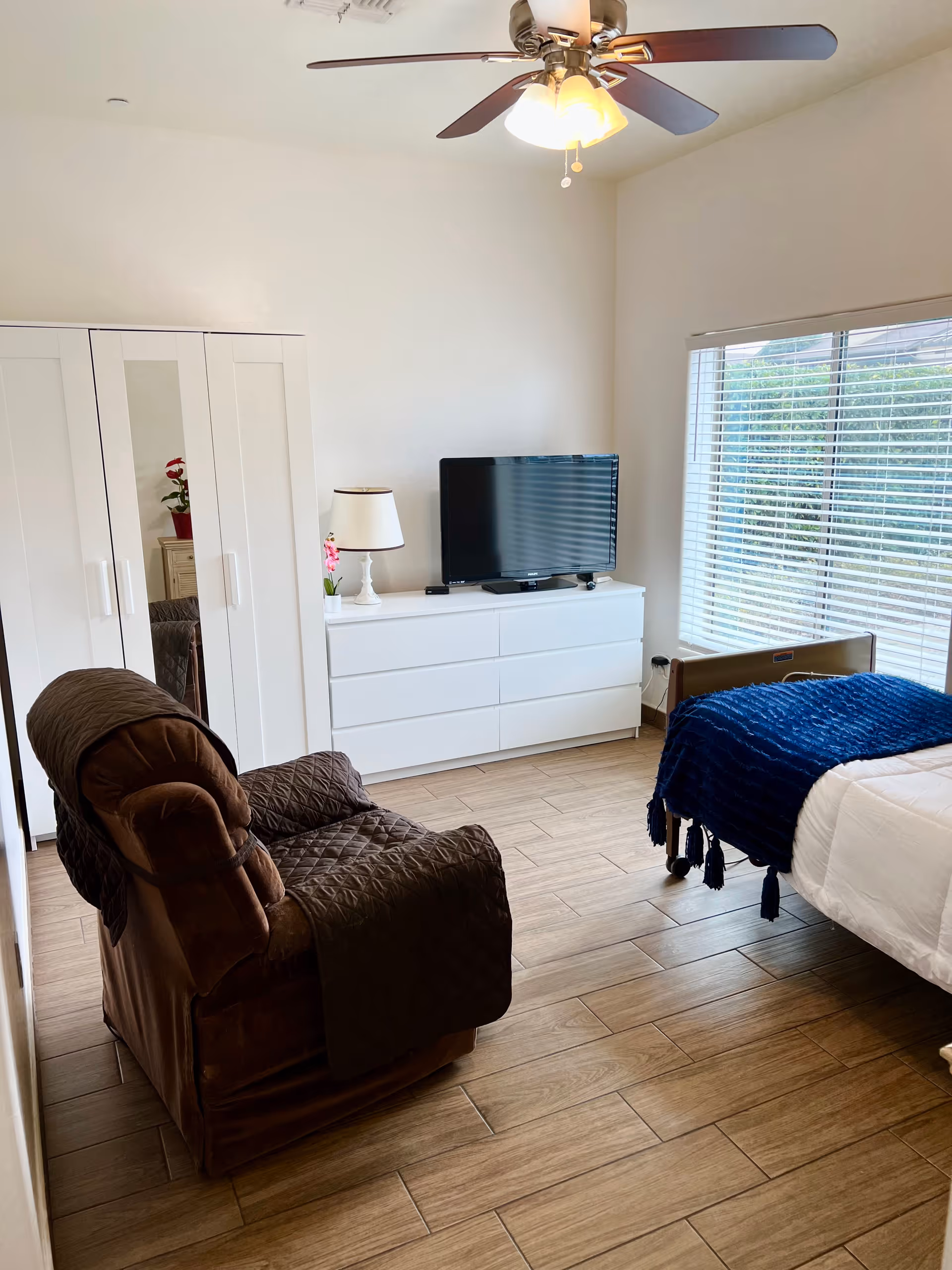 A bright bedroom with a brown recliner chair covered with a quilted throw, a white dresser with a flat-screen TV and a lamp on top, a white wardrobe with mirrored doors, a bed with white bedding and a blue throw blanket, and a large window with blinds letting in natural light. The floor is wood-style tile and there is a ceiling fan with lights.