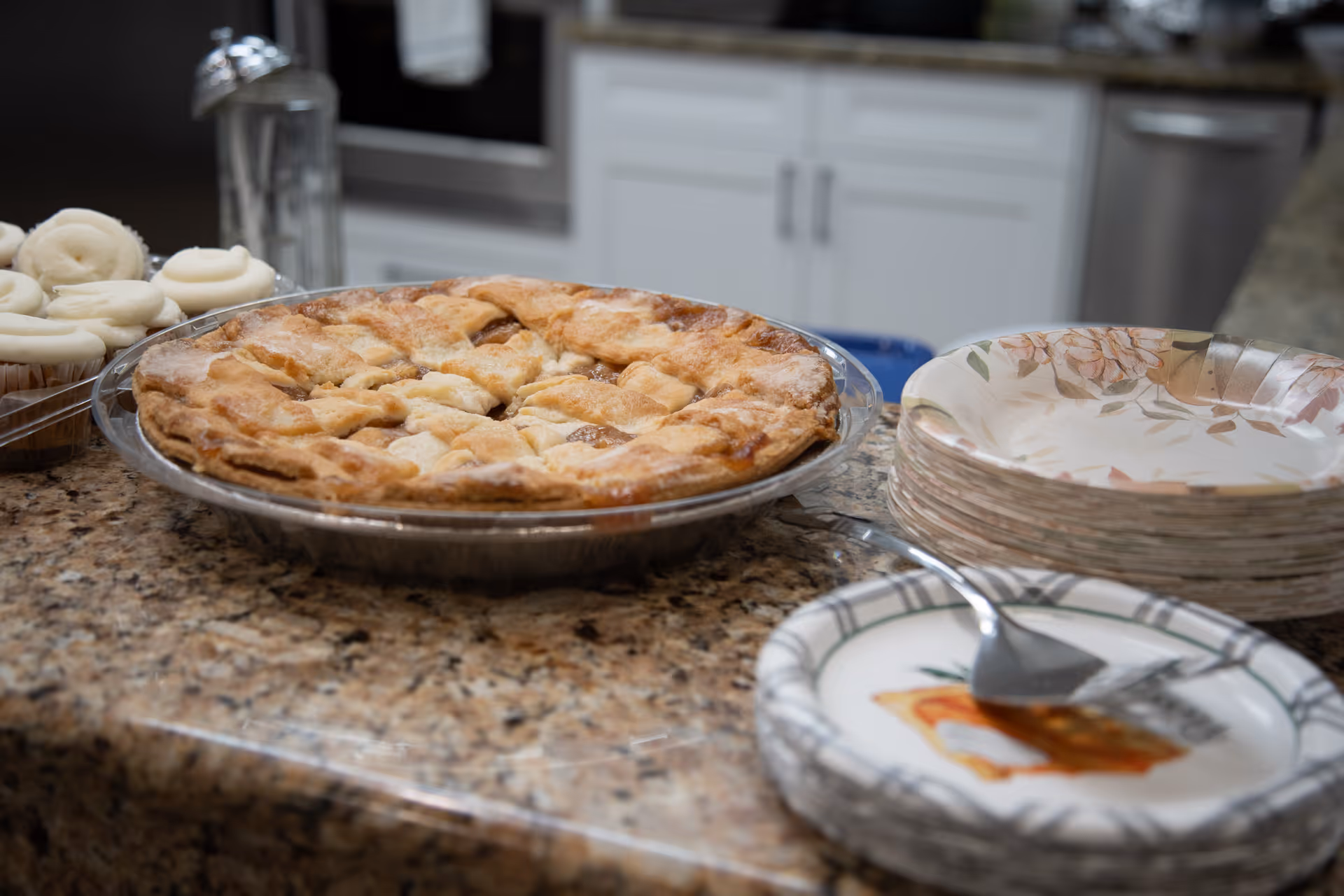 A freshly baked pie and cupcakes on a granite kitchen counter next to stacks of paper plates and a serving spatula.