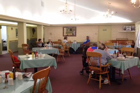 A communal dining room with multiple tables covered in green tablecloths and elderly residents seated and chatting.