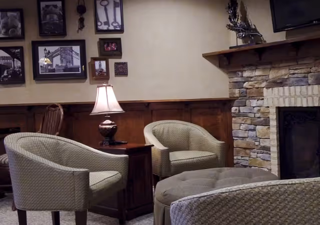 A cozy living room area with three cushioned armchairs arranged around a round ottoman. There is a wooden side table with a lamp on it between two of the chairs. The room features a stone fireplace on the right side and a wall decorated with framed pictures and keys.