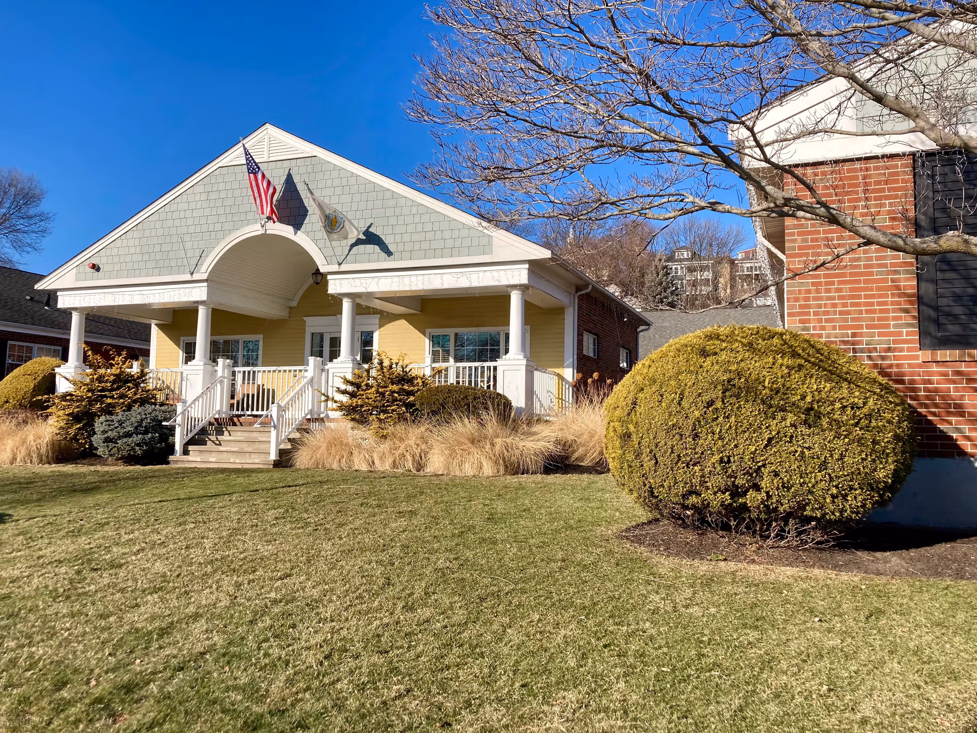 Exterior view of a single-story building with a porch, white columns, and two flags flying above the entrance. The building has light green shingles on the gable and yellow siding below. There is a well-maintained lawn and trimmed bushes in front, with a clear blue sky and leafless tree branches overhead.