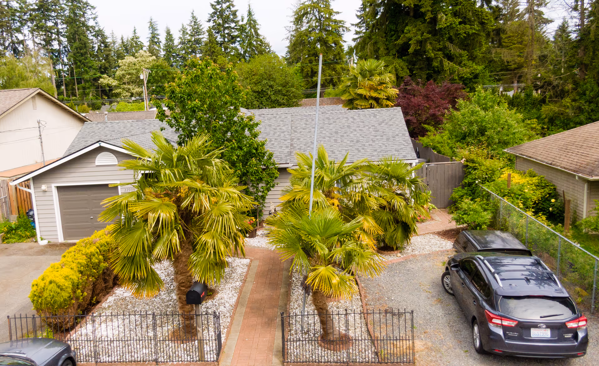 Front exterior of a single-story house with palm trees along a brick walkway, gravel driveway, and parked cars.