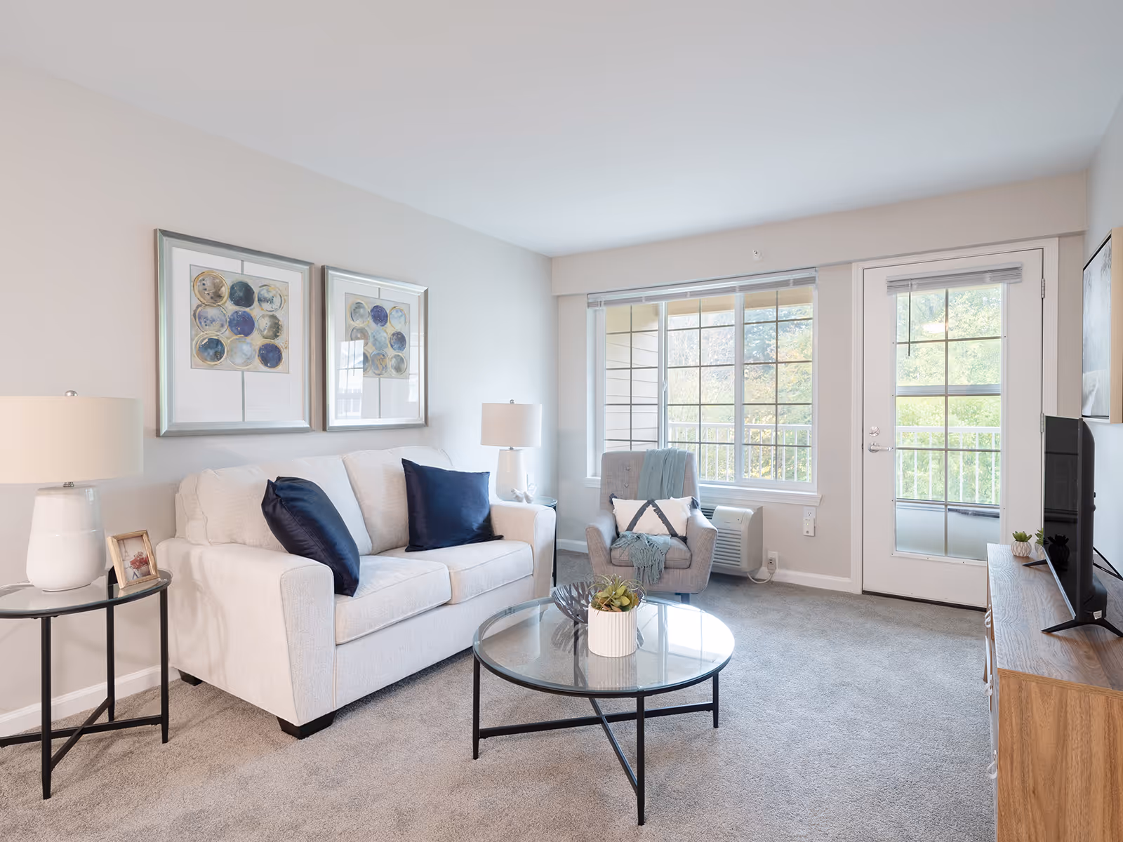 A bright living room with a light gray sofa adorned with dark blue pillows, a gray armchair with a white and blue pillow and a blue throw, a round glass coffee table with a small plant, two framed abstract artworks on the wall, a side table with a white lamp and a small photo frame, a large window and a glass door leading to a balcony with greenery outside, and a wooden TV stand with a flat-screen TV.
