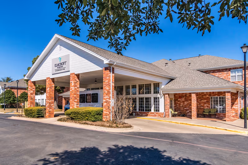 Front entrance of a red-brick assisted living building with a covered porte-cochère, shrubs, and a parked van under a clear blue sky.
