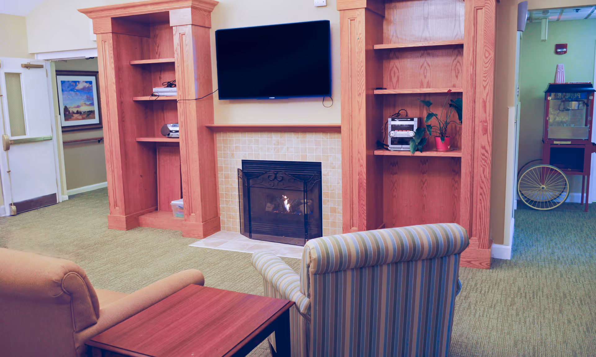 A cozy common area with a fireplace surrounded by wooden built-in shelves, a flat-screen TV mounted above the fireplace, and two armchairs facing the fireplace. There is a small wooden side table between the chairs. In the background, a popcorn machine and a hallway with a framed picture on the wall are visible.