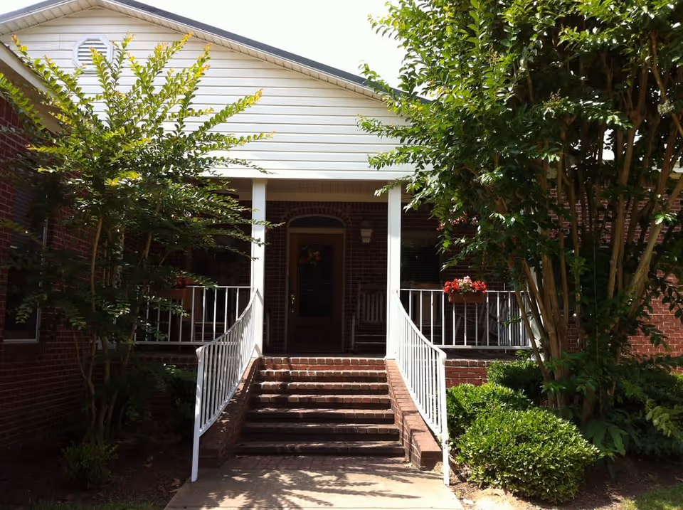 Front entrance of a brick building with a white porch roof, white railings, and steps leading up to a wooden door. There are green bushes and trees on either side of the entrance, with a flower box containing red flowers on the porch railing.