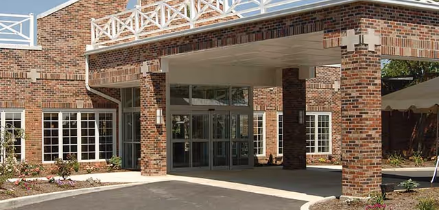 Entrance of a brick building with large glass doors under a covered drop-off area supported by brick pillars, surrounded by landscaped flower beds and greenery.
