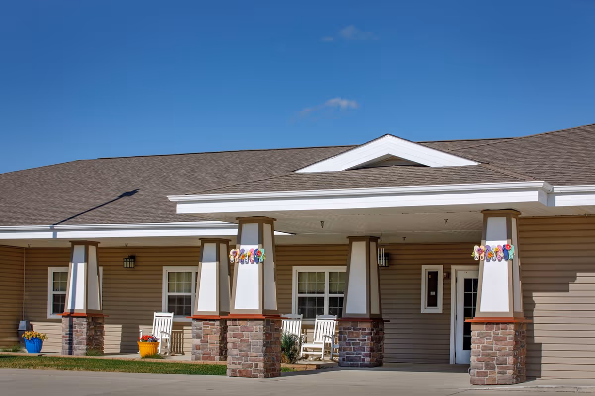 Front entrance of an assisted living facility with a covered canopy supported by stone pillars, rocking chairs, and colorful planters under a blue sky.