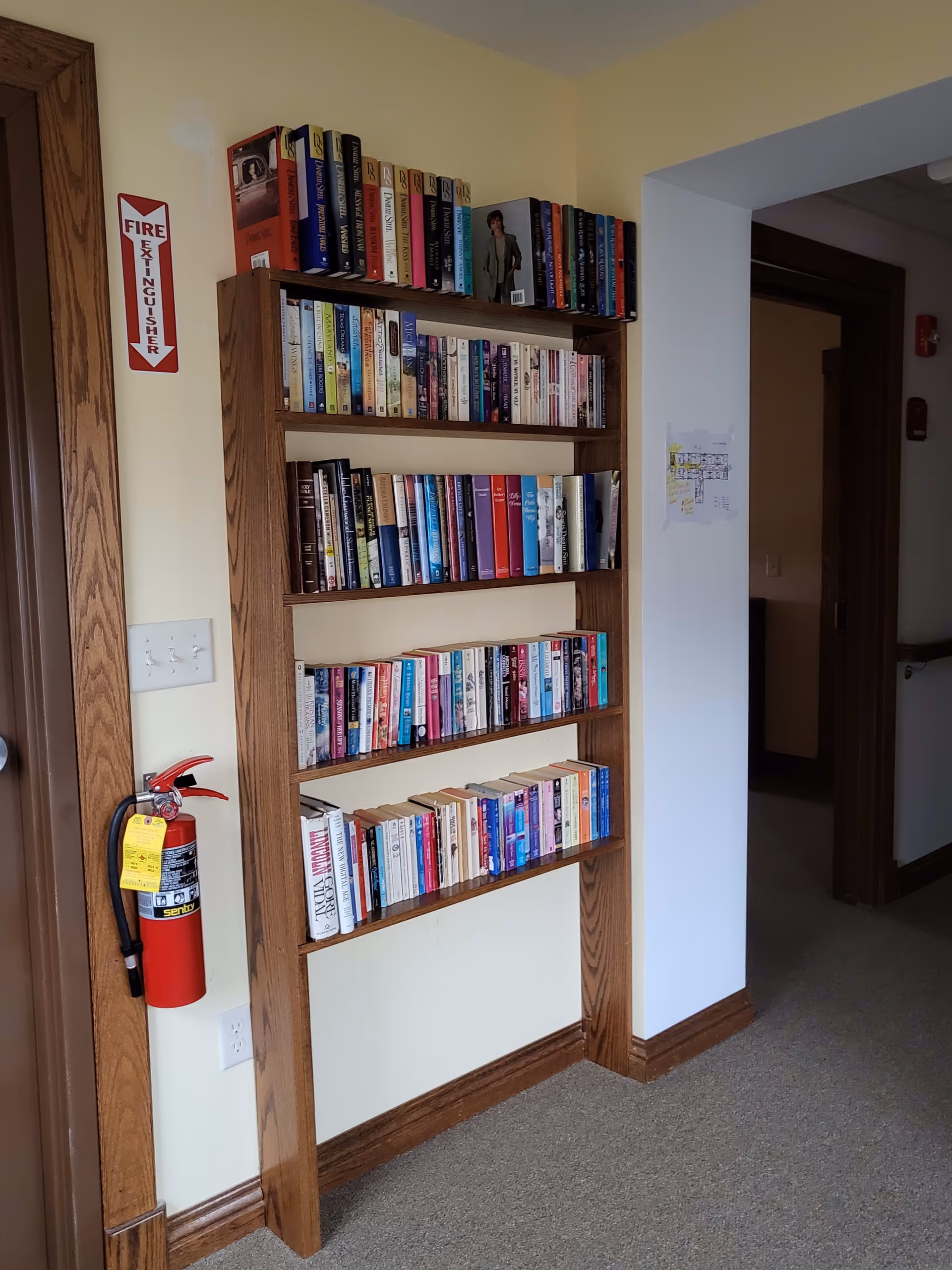 Wooden built-in bookshelf filled with books in a hallway next to a fire extinguisher and an open doorway.