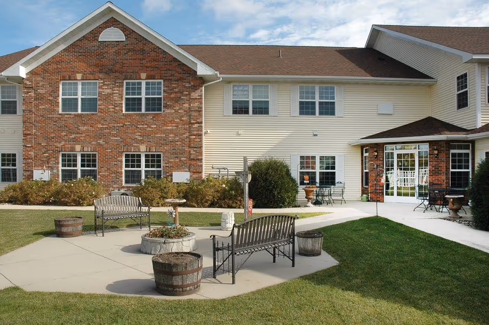 Outdoor courtyard with benches, planters, and seating in front of a two-story brick-and-siding senior living building.