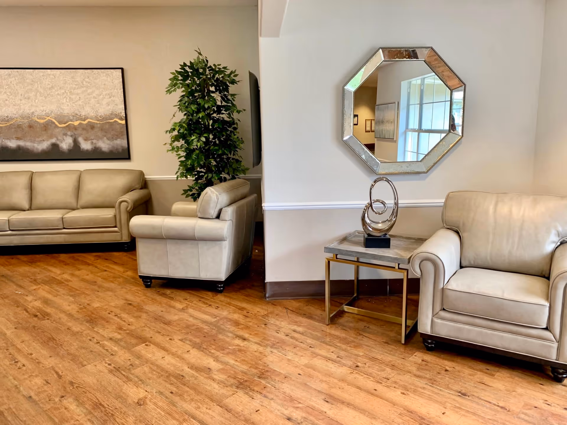 A cozy living room area with beige leather sofas and armchairs, a wooden floor, a tall green plant, a side table with a decorative sculpture, a large abstract painting on the wall, and an octagonal mirror reflecting a window and part of the room.